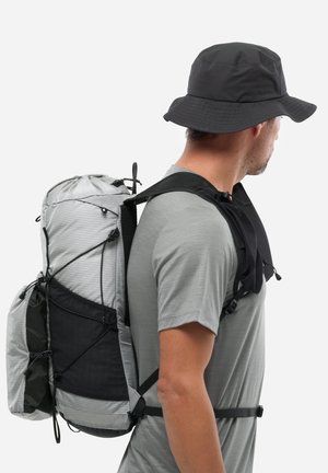 Homme portant une chemise grise et un chapeau seau noir avec un grand sac à dos gris, vu de profil sur un fond blanc uni.