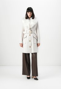 Vrouw in een witte blestriemjas met een bontkraag, bruine wijde broek en zwarte blokhak schoenen, staande voor een witte achtergrond.