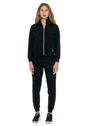 Une femme porte une veste noire zippée assortie à un pantalon de jogging, ainsi que des baskets noires, sur un fond blanc uni.