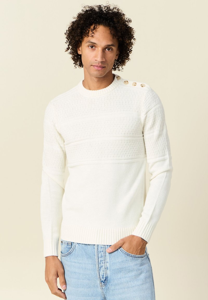 Maglione a maglia crema con strisce orizzontali testurizzate e tre bottoni sulla spalla. Abbinato a jeans di denim azzurri chiari.