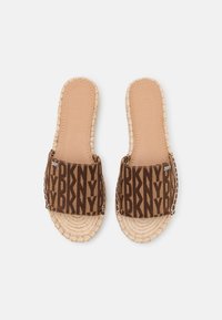 Brown slide sandals featuring a DKNY logo pattern on fabric straps, with a braided jute sole and soft leather inner lining.