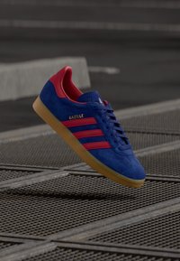 Ténis Adidas Gazelle azul e vermelho com sola de borracha, flutuando acima de uma superfície de grelha metálica ao ar livre.