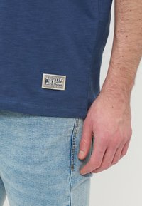 Εγγύς πλάνο του χεριού ενός ατόμου που ακουμπά στο πλάι του, φορώντας ανοιχτό μπλε τζιν και μπλε πουκάμισο με ετικέτα Pier One.