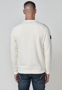Sudadera blanca texturizada con un patrón de cuadrícula, puños y cintura ajustados. Presenta una etiqueta negra en la manga izquierda. Combinada con jeans azules.