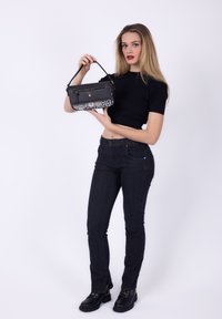 Kobieta w czarnym topie crop i dżinsach, trzymająca czarną torbę na ramię z wzorzystym dnem, stojąca na tle białym.