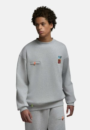 Jeune personne portant un sweat-shirt gris et un pantalon assorti avec de petites pièces colorées et des détails orange sur les poches.