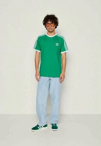 Ung mann med krøllete hår, iført oransje tonede briller, grønn Adidas t-skjorte, lyseblå vide jeans og grønne Adidas joggesko, som står foran en ensfarget bakgrunn.