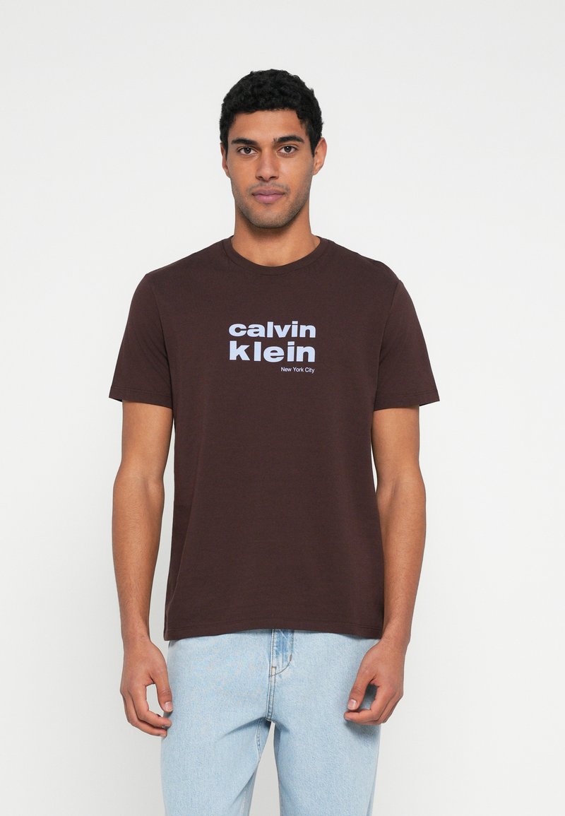 Tricou maro din bumbac cu decolteu rotund și mâneci scurte, având inscripționate pe față cu litere albe mari textele „calvin klein” și „New York City”.