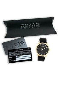 Montre noire avec des accents dorés, cadran rond, chiffres arabes, bracelet en cuir. Comprend un emballage noir et des cartes de garantie.