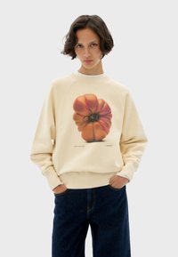 Cream-colored sweatshirt featuring an orange and green pumpkin graphic. Fabric is soft, with ribbed cuffs and a relaxed fit. Worn with dark jeans.