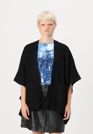 Poncho nero lavorato a maglia con maniche ampie e due tasche frontali, indossato sopra un top blu con paillettes; abbinato a una gonna di pelle nera.