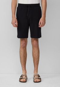 Schwarze gewebte Shorts mit einem elastischen Bund und Kordelzug, im entspannten Schnitt und hemdenlang bis über das Knie. Getragen mit braunen Sandalen.