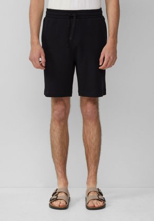 Shorts noirs en tissu tissé avec une taille élastique et un cordon de serrage, dotés d'une coupe décontractée et arrivant au-dessus du genou. Portés avec des sandales marron.