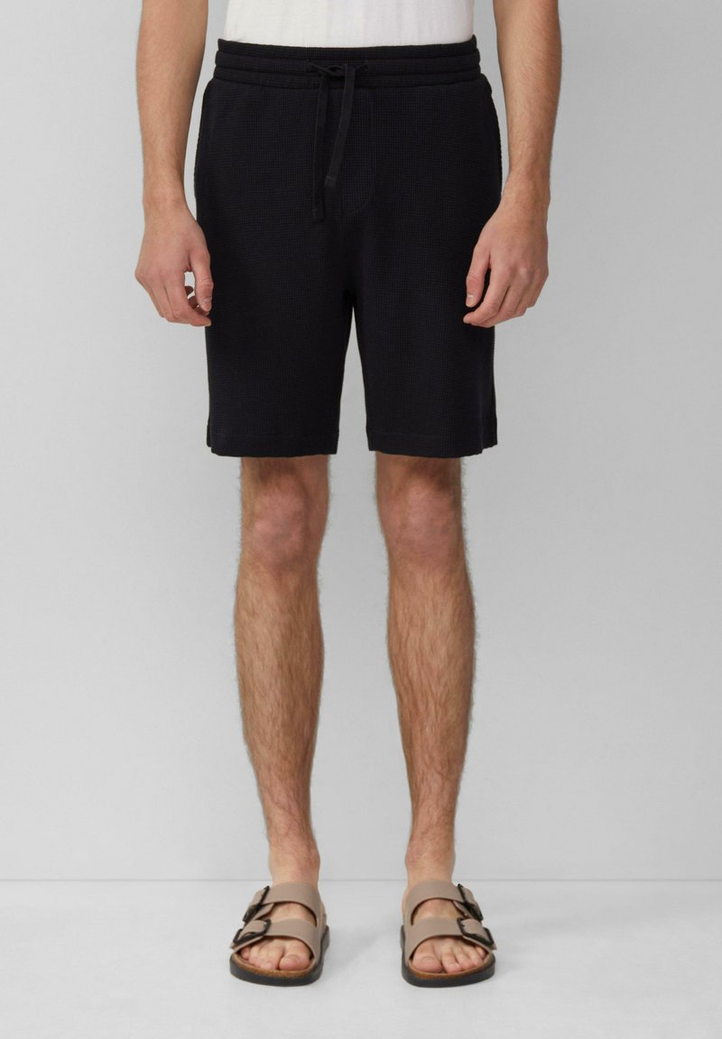Schwarze gewebte Shorts mit einem elastischen Bund und Kordelzug, im entspannten Schnitt und hemdenlang bis über das Knie. Getragen mit braunen Sandalen.