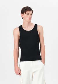 Sort ærmeløs tanktop lavet af glat stof, med rund halsudskæring og en tætsiddende pasform, parret med lyse bukser.