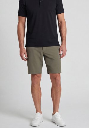 Mannelijk model met een zwart poloshirt met korte mouwen, olijfgroene shorts en witte sneakers, staand tegen een effen lichtgrijze achtergrond.