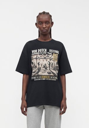 Femme aux cheveux tressés portant un t-shirt noir oversize avec un imprimé vintage de festival de musique et un pantalon gris, debout sur un fond uni.
