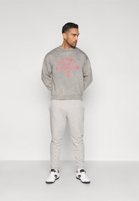 Pelēka sporta džemperis ar sarkanu uzrakstu "ASV vīriešu nacionālās futbola komandas" grafiku. Pelēkas sporta bikses. Baltas sporta kurpes ar melniem akcentiem. Stāvoša poza.