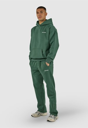 Uomo in piedi che indossa una felpa con cappuccio verde scuro e pantaloni della tuta abbinati con sneakers bianche su sfondo grigio chiaro.