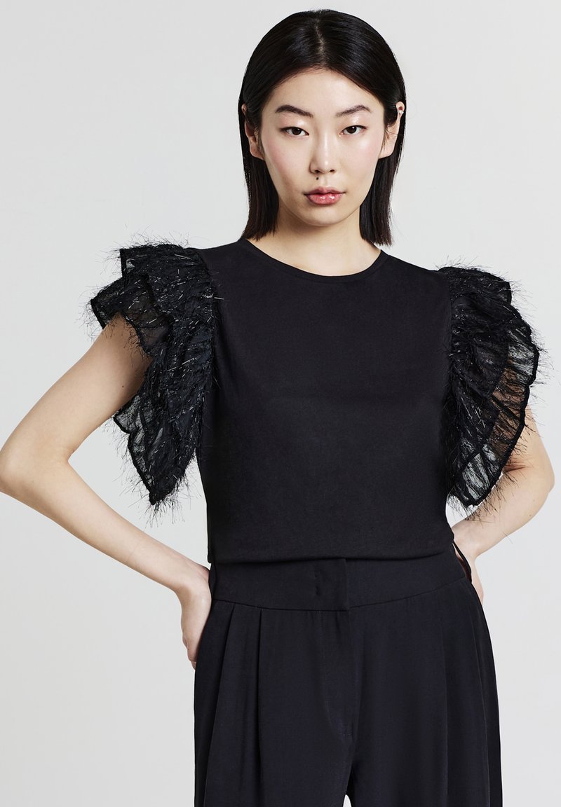 Giovane donna con capelli neri lisci, che indossa un top nero senza maniche con maniche in tulle trasparente arricciate e pantaloni neri, con le mani sui fianchi.