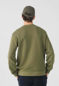 Hombre con sudadera verde oliva y gorra a juego, de espaldas, con las manos en los bolsillos frente a un fondo claro y liso.