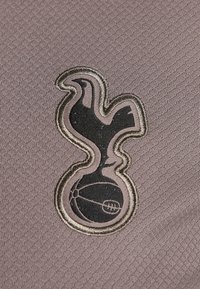 La tela gris presenta un logotipo bordado en negro de un pájaro con una base esférica, delineado en dorado, exhibiendo texturas suaves y líneas limpias.