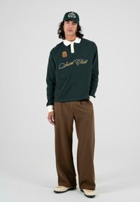 Groene lange mouwen polo met witte kraag en borduring "Social Club", gecombineerd met bruine wijde broek en crèmekleurige sneakers.