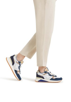 Zapatillas deportivas en azul marino y beige con acentos blancos, diseño con cordones y suelas texturizadas. La parte superior presenta materiales mezclados y un patrón geométrico.