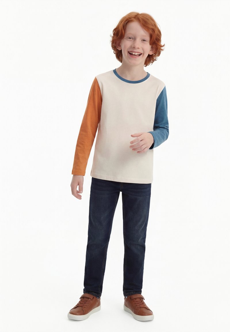 Garçon aux cheveux roux souriant, portant une chemise à manches longues avec des manches orange et bleues, un jean foncé et des baskets marron, debout devant un fond blanc.