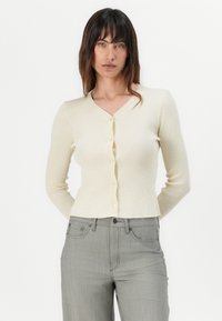 Cardigan a coste crema con scollo a V, vestibilità corta e chiusura frontale con sei bottoni. Maniche lunghe e texture liscia e morbida.