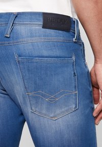 Jeans in denim blu con una tasca posteriore caratterizzata da un motivo cucito di colore marrone chiaro. Un'etichetta in pelle nera sulla cintura mostra la scritta "REPLAY".