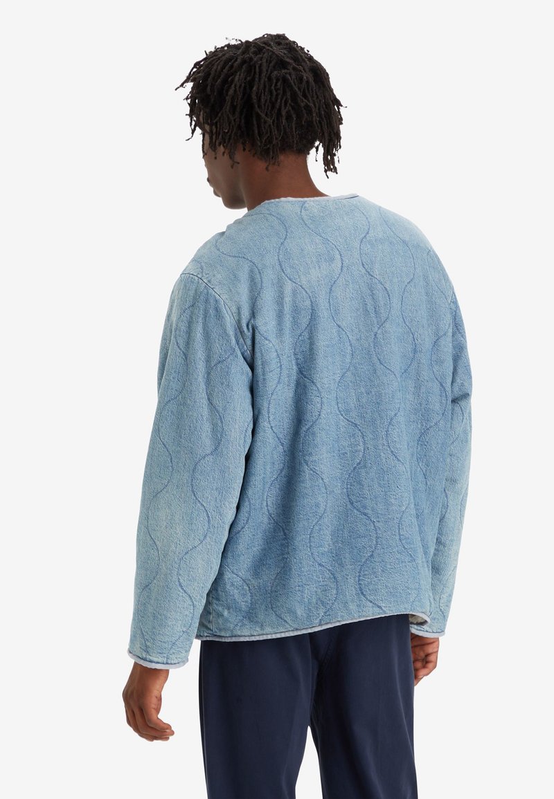 Chaqueta de mezclilla acolchada de color azul claro con una textura ondulada, cuello redondo y mangas largas. Pantalones oscuros son visibles en el borde inferior.