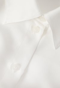 Chemise blanche avec un col classique, une texture lisse et deux petits boutons ronds. Le tissu semble léger avec un motif de tissage subtil.
