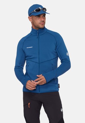 Uomo che indossa una giacca blu con zip, un cappellino blu coordinato con occhiali da sole sopra, e pantaloni neri con loghi visibili.