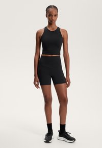 Femme avec des cheveux tressés portant un crop top noir sans manches, des shorts de vélo taille haute, des chaussettes noires et des chaussures de course noires, se tenant droite.