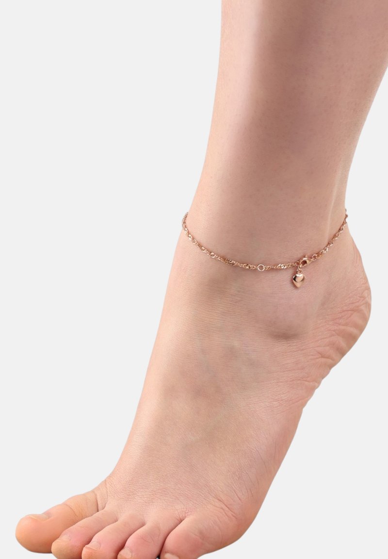 Primer plano de un pie descalzo que lleva un delicado tobillo de oro rosa con un pequeño colgante de corazón sobre un fondo blanco liso.