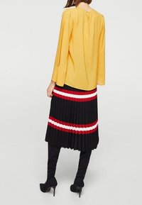 Blusa gialla con design a pieghe sul retro, abbinata a una gonna nera plissettata con motivi a righe rosse e bianche. Stivali alti neri completano il look.