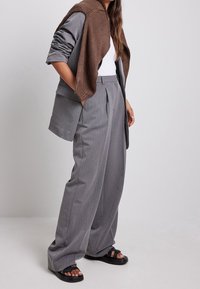 Pantalon gris à jambes larges avec des plis sur le devant, associé à un t-shirt blanc et un châle en tricot marron. Le look est complété par des sandales noires.