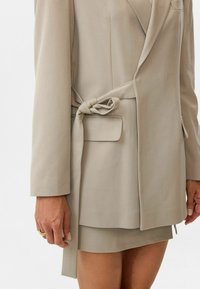 Beige blazer met een gestrikt riem, scherpe revers en een voorzak. De stof is glad met een getailleerde pasvorm, die een bijpassende rok aanvult.