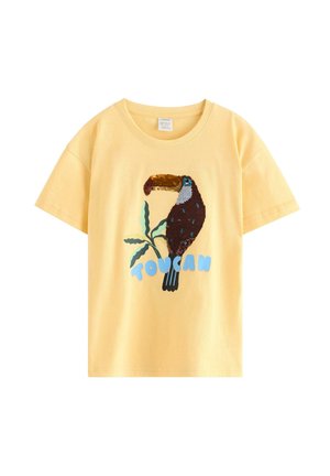 Ung dreng iført en stråhat, gul t-shirt med en tucan-grafik, brune shorts, strømper med tucan-mønster og brune velcro-sneakers.