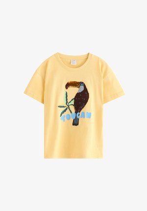 Ung dreng iført en stråhat, gul t-shirt med en tucan-grafik, brune shorts, strømper med tucan-mønster og brune velcro-sneakers.