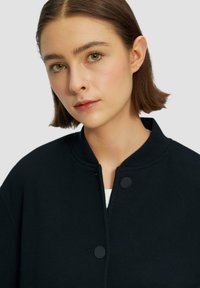 Veste noire texturée avec un col rond, présentant deux boutons à l'avant et une coupe décontractée. Tissu lisse avec un motif subtil.