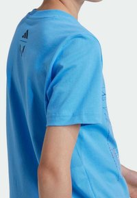 Persona que lleva una camiseta de manga corta azul brillante con logotipos negros cerca de la parte superior de la espalda, mostrada de lado y por detrás.