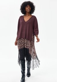 Bordowy sweter z dekoltem w serek, zestawiony z przezroczystą spódnicą w panterkę i czarnymi koronkowymi detalami; modelka ma na sobie czarne kozaki za kolano.