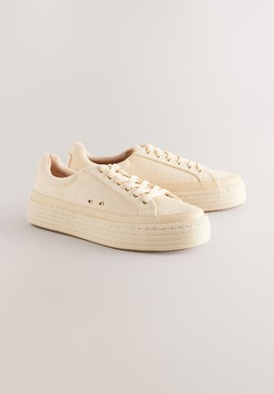 Bézs vászon sportcipők struktúrált felsőrészsel, kerek orral és vastag, kötélhatású talppal. Lapos fűzők és aranyszínű fémfülek jellemzik.