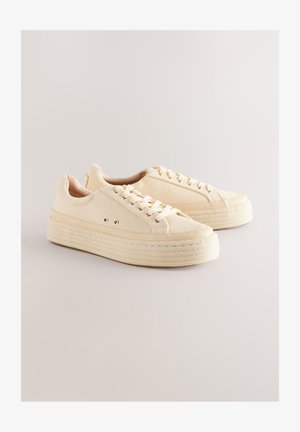 Bézs vászon sportcipők struktúrált felsőrészsel, kerek orral és vastag, kötélhatású talppal. Lapos fűzők és aranyszínű fémfülek jellemzik.