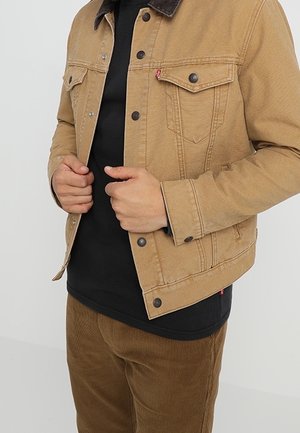 Osoba ubrana w jasnobrązową kurtkę dżinsową na czarnej koszulce i brązowe sztruksowe spodnie, poprawiająca przód kurtki obiema rękami.