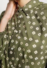 Camicia verde con motivo, colletto, caratterizzata da disegni bianchi a forma quadrata e chiusura con bottoni. Il tessuto appare morbido e testurizzato.