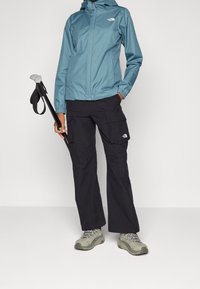 Vattentålig blå jacka med huva, i kombination med svarta cargobyxor och grå vandringsskor. Inkluderar en trekkingstav med svart handtag.