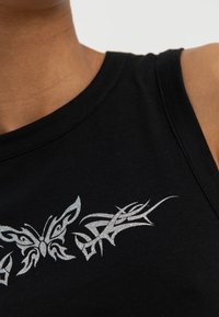 Crni top bez rukava s motivom leptira i plemenskim dizajnom u srebrnom sjaju. Mekani pamuk s glatkom teksturom i širokim naramenicama.